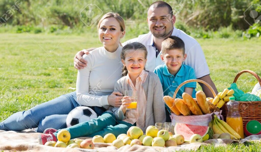 una foto de una familia sonriente disfrutando de un dia soleado en un campo verde con una canasta de picnic y jugando a la pelota